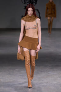 Model walks the runway in a tan fur-neck collar, a chain-bra top, and a short tan skirt with over-the-knee boots.