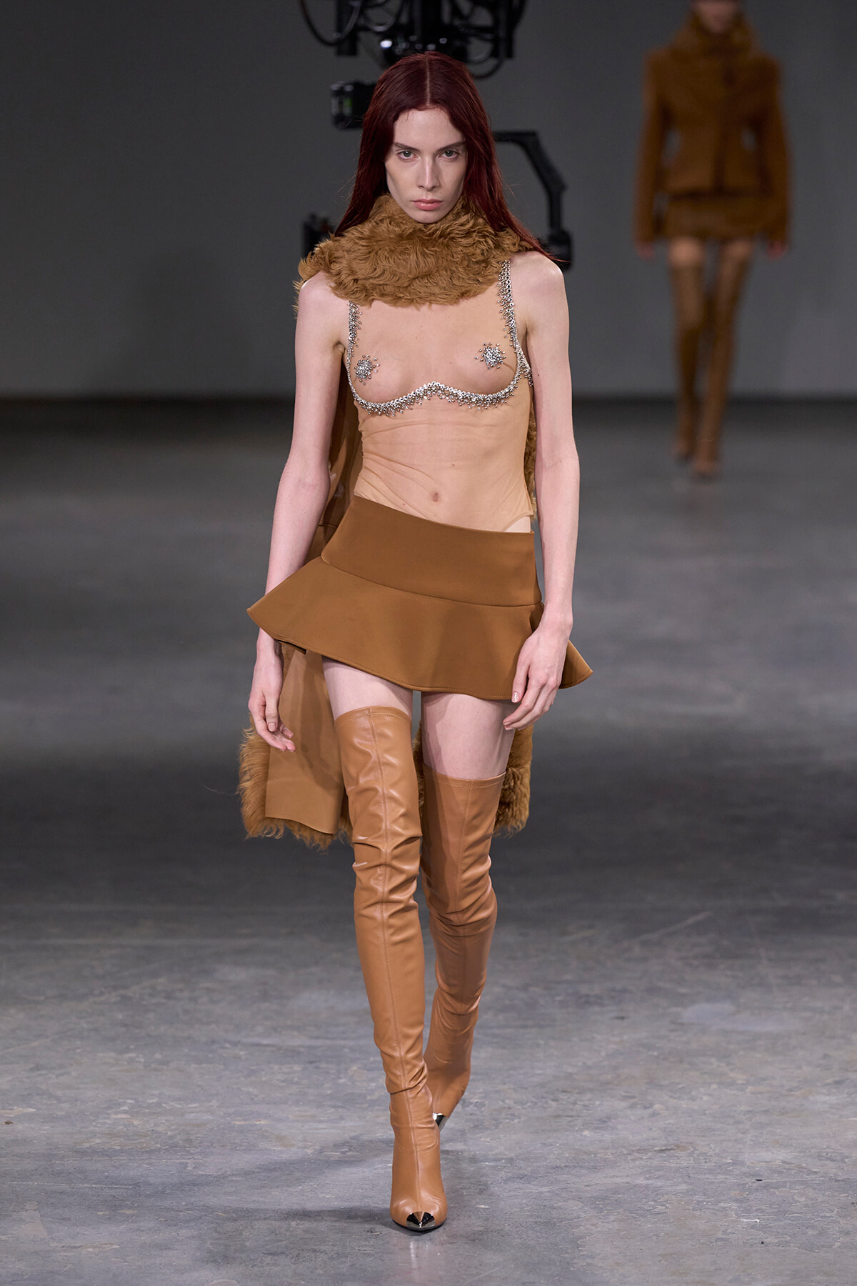 Model walks the runway in a tan fur-neck collar, a chain-bra top, and a short tan skirt with over-the-knee boots.