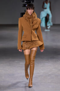 Model walking a fashion runway in a camel blazer, matching mini skirt with beige faux-fur trim and over-the-knee boots.
