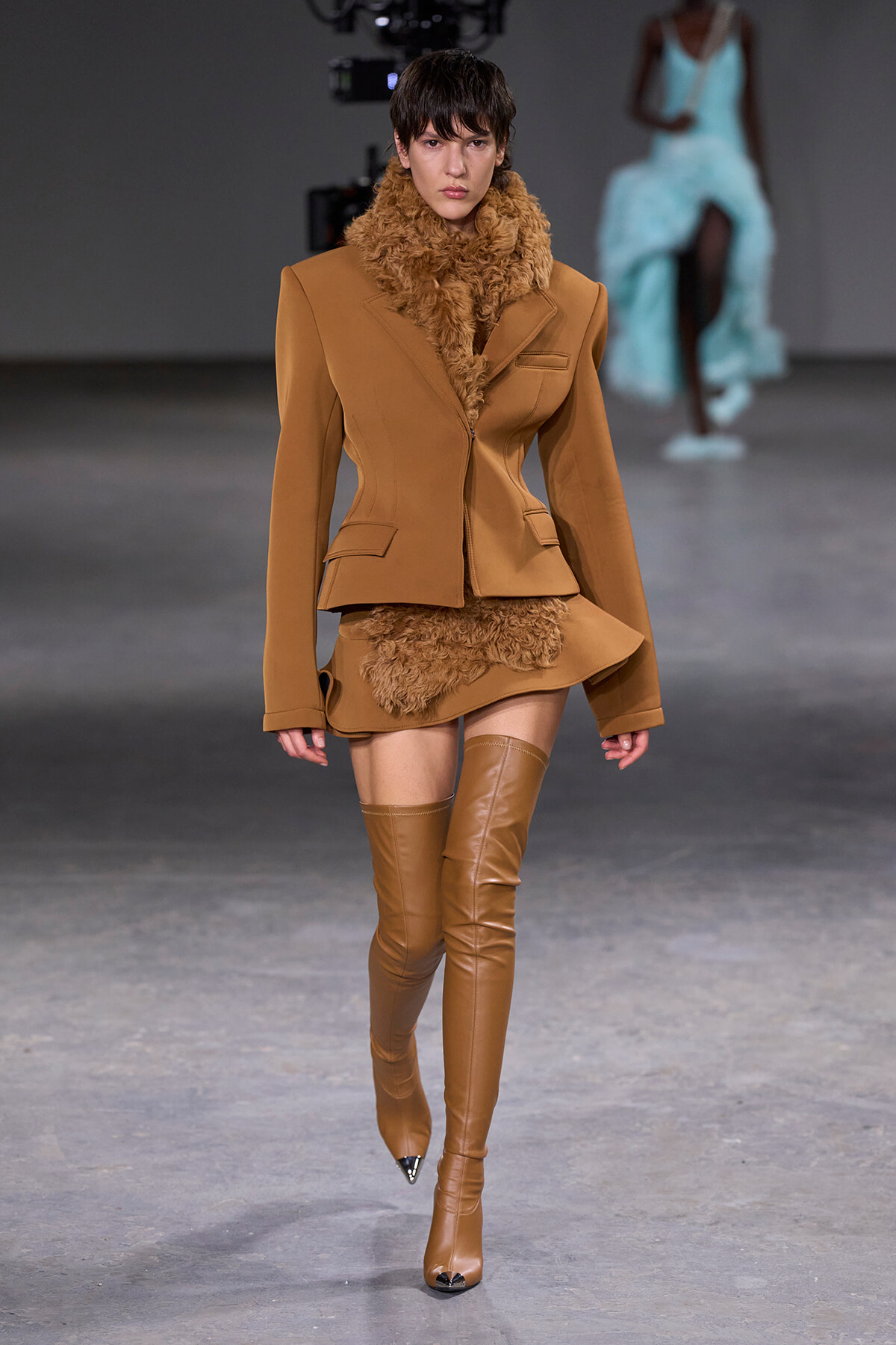 Model walking a fashion runway in a camel blazer, matching mini skirt with beige faux-fur trim and over-the-knee boots.