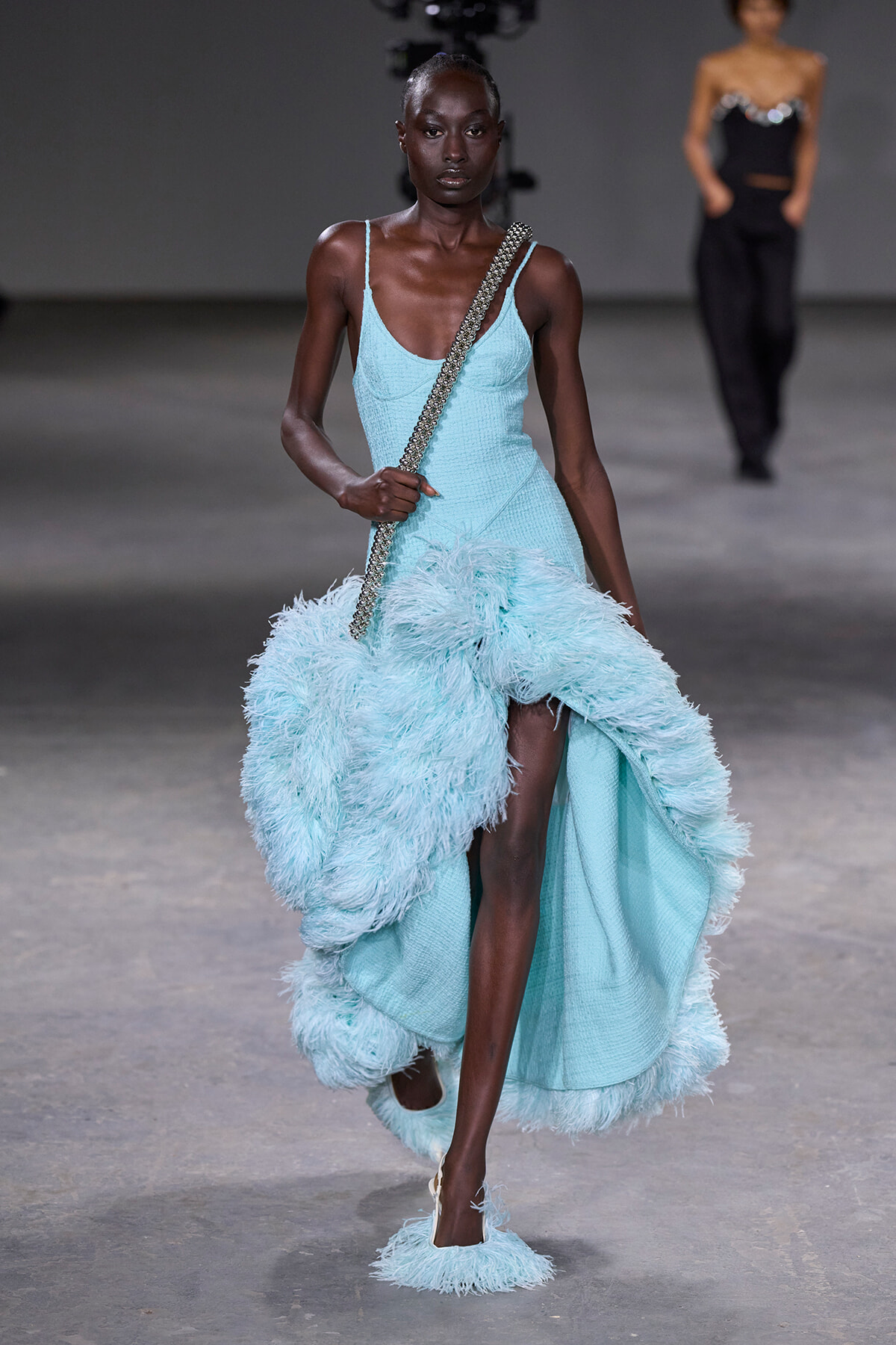 Model walks the runway in a pale turquoise gown with a voluminous feathered train and a sparkling silver chain crossbody bag.