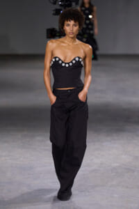 Model walking a fashion runway in a black strapless top with crystal embellishments and black trousers.