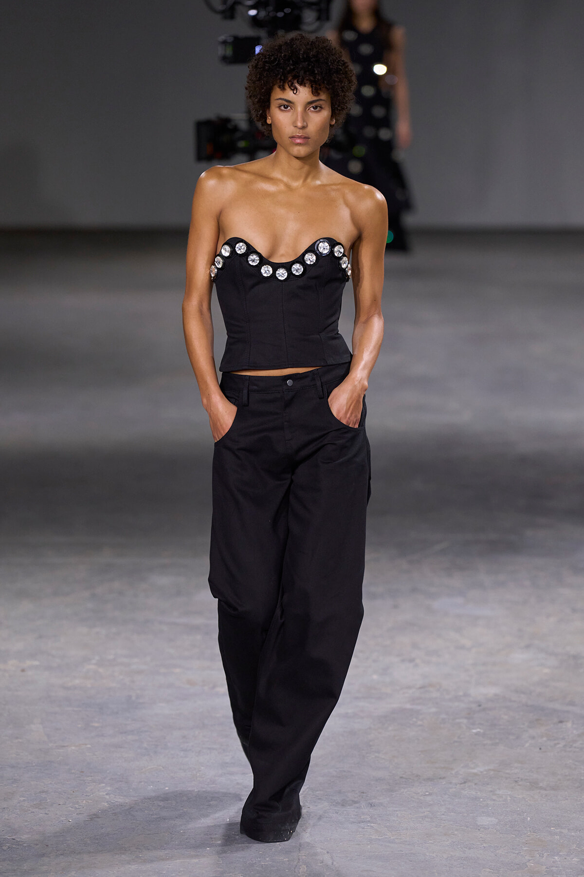 Model walking a fashion runway in a black strapless top with crystal embellishments and black trousers.