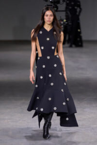 Model walking a runway in a black, sleeveless dress with crystal circle embellishments and cutouts on the sides.