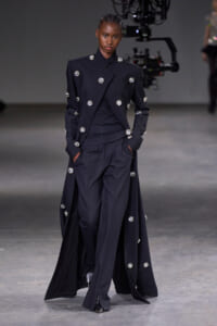 Full-length view of a model walking the runway in a navy double-breasted coat with large crystal buttons and matching trousers.