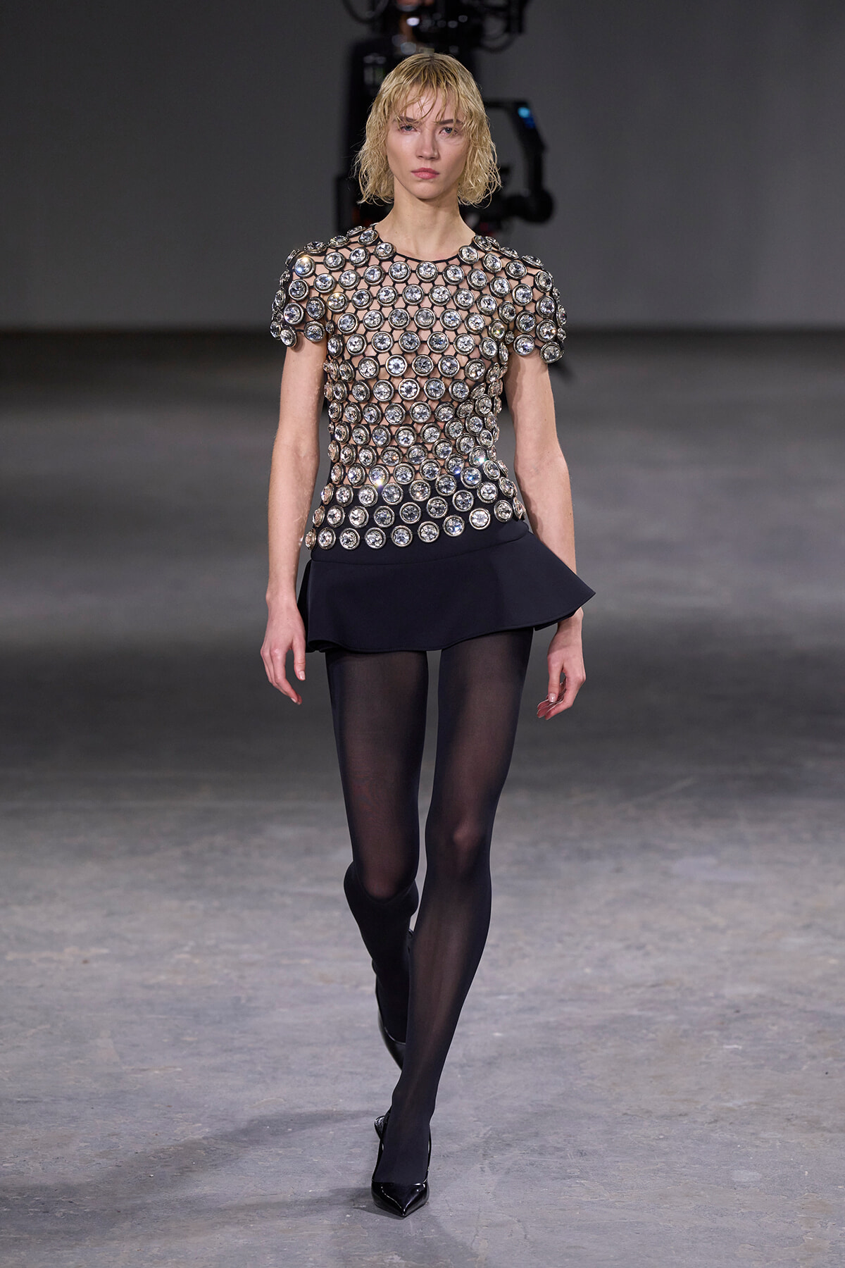 Model walking the runway in a silver, circular-disc embellished sheer top with a short black skirt and black tights.