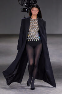 Model struts a runway in an oversized black blazer over a top covered with circular silver discs, paired with black tights and heels.