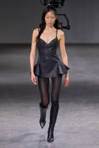 Model walking a fashion runway in a black leather corset-style top with a peplum skirt and black tights and boots.