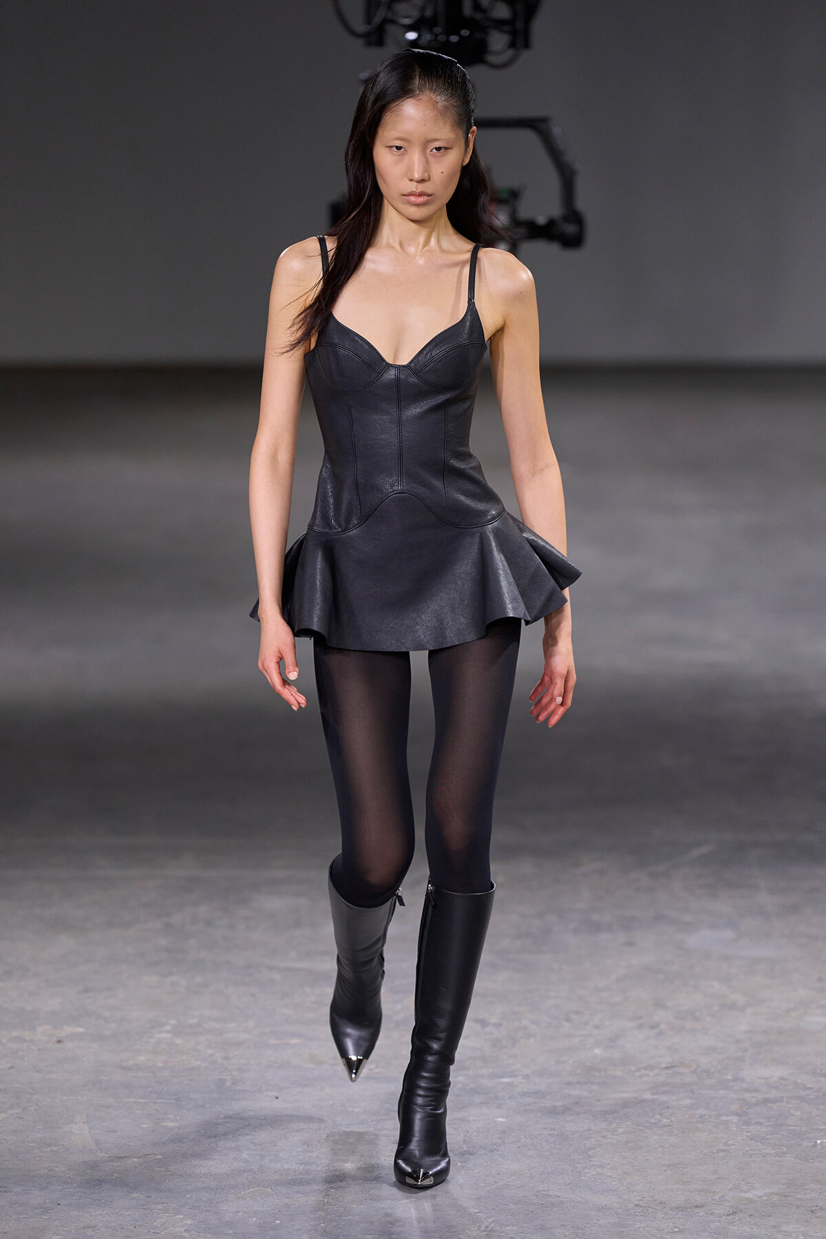 Model walking a fashion runway in a black leather corset-style top with a peplum skirt and black tights and boots.