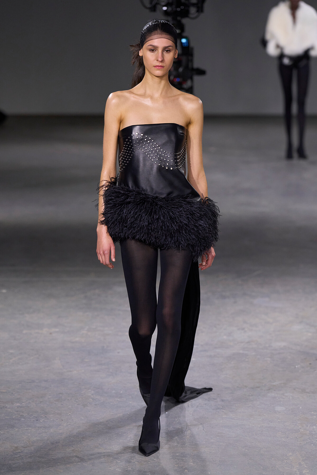 Model walks a runway in a black strapless top with studded silver embellishment and a dense black feathered hem, paired with opaque tights and pointed black heels.