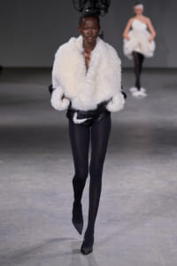 Model walks the runway in a voluminous white shearling jacket over black leggings and pointed black heels.