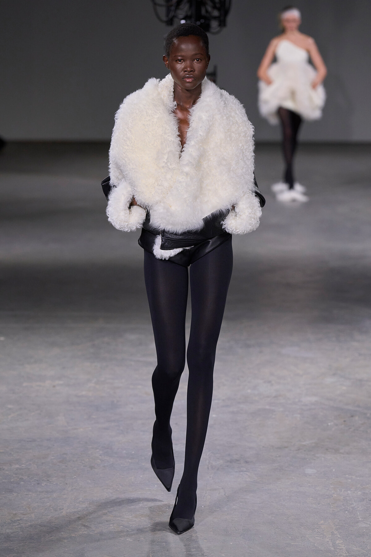 Model walks the runway in a voluminous white shearling jacket over black leggings and pointed black heels.