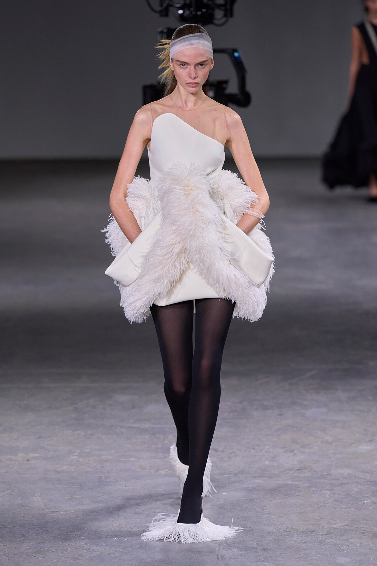 Model walks the runway in a white one-shoulder dress with a voluminous feathered skirt and black tights.