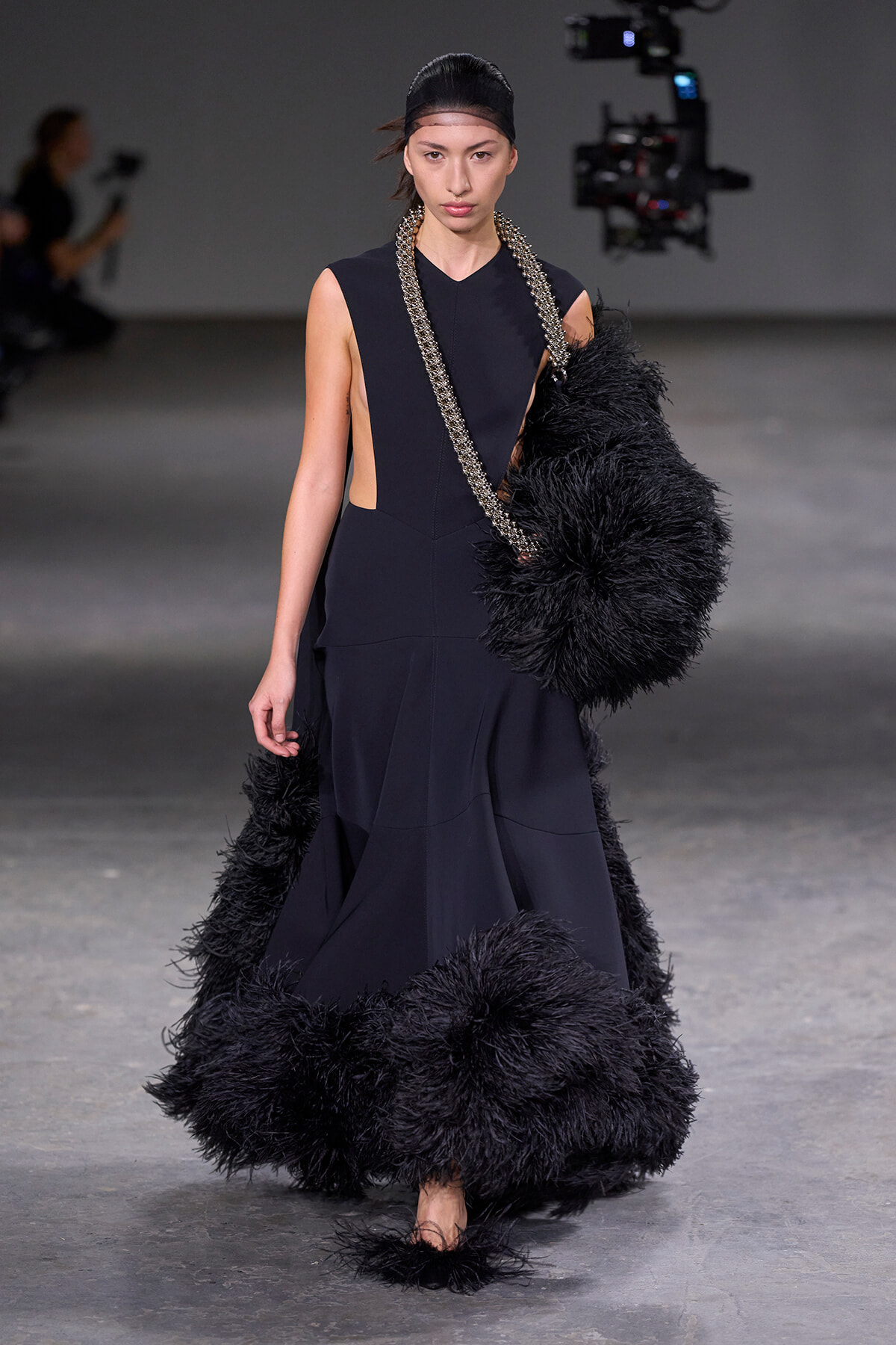 Model walking a runway in a sleeveless black gown with cutouts, holding a large black feathered scarf/boa.