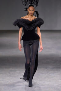 Model walking a fashion runway in a black off‑the‑shoulder top with a voluminous feathered collar and black leggings