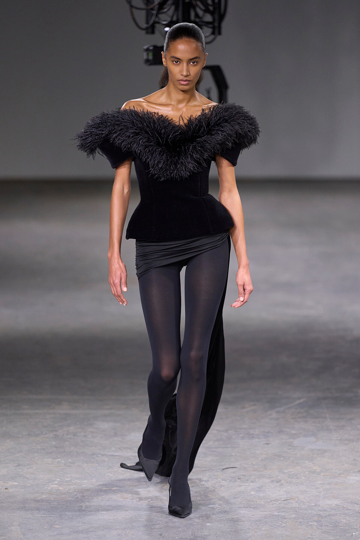 Model walking a fashion runway in a black off‑the‑shoulder top with a voluminous feathered collar and black leggings