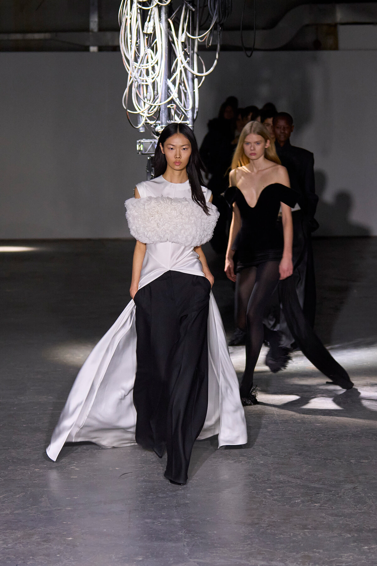 Model on a runway wearing a white satin gown with a voluminous textured bodice and black trousers; overhead cables loom above.
