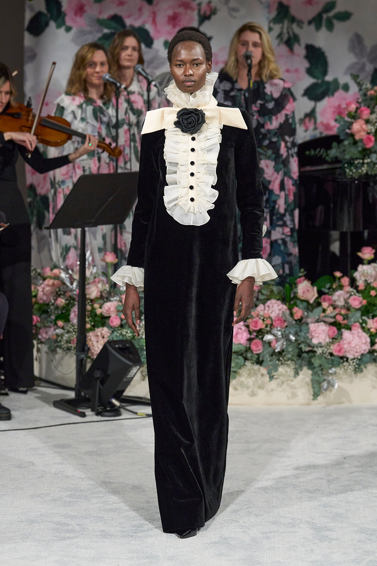 Model wearing a black velvet gown with white ruffled bib and large neck bow on a floral-themed runway; musicians visible in the background.