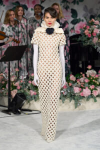 Model on a runway wearing a full-length white lattice gown with long gloves and a black rose neck ornament, floral backdrop in background.