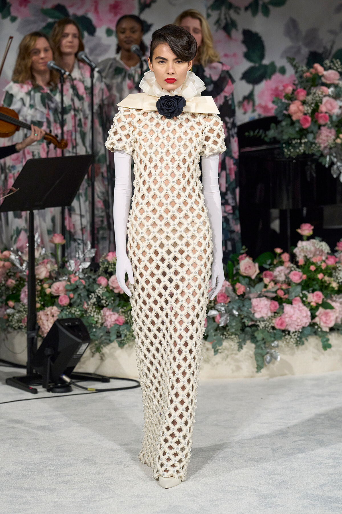 Model on a runway wearing a full-length white lattice gown with long gloves and a black rose neck ornament, floral backdrop in background.