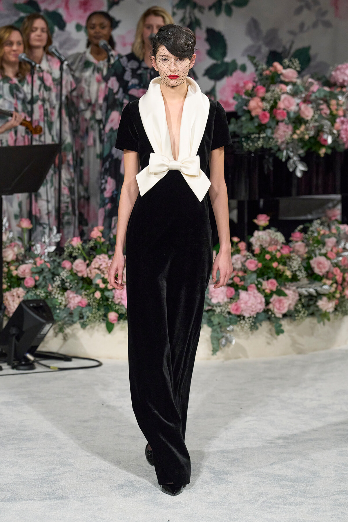 Model on a floral-themed runway wearing a black velvet jumpsuit with a large white satin bow at the waist and a sheer net veil over her face.