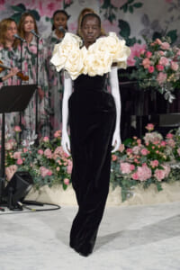 Runway model walks wearing a black velvet gown with an oversized cream ruffled collar, floral backdrop behind and pink arrangements to sides