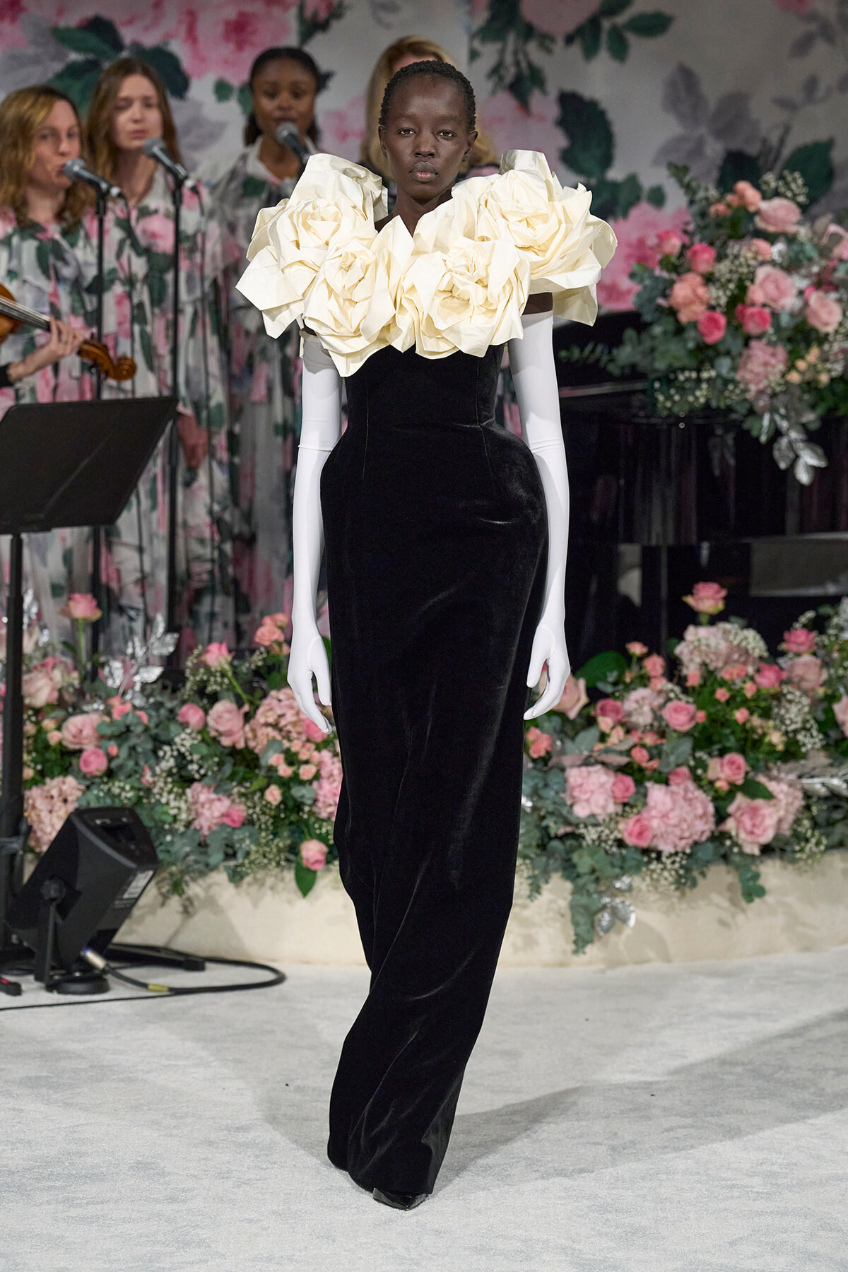 Runway model walks wearing a black velvet gown with an oversized cream ruffled collar, floral backdrop behind and pink arrangements to sides
