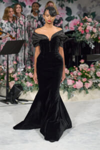 Model walking the runway in a black off-the-shoulder velvet gown with a small veil over her face, floral backdrop behind her.