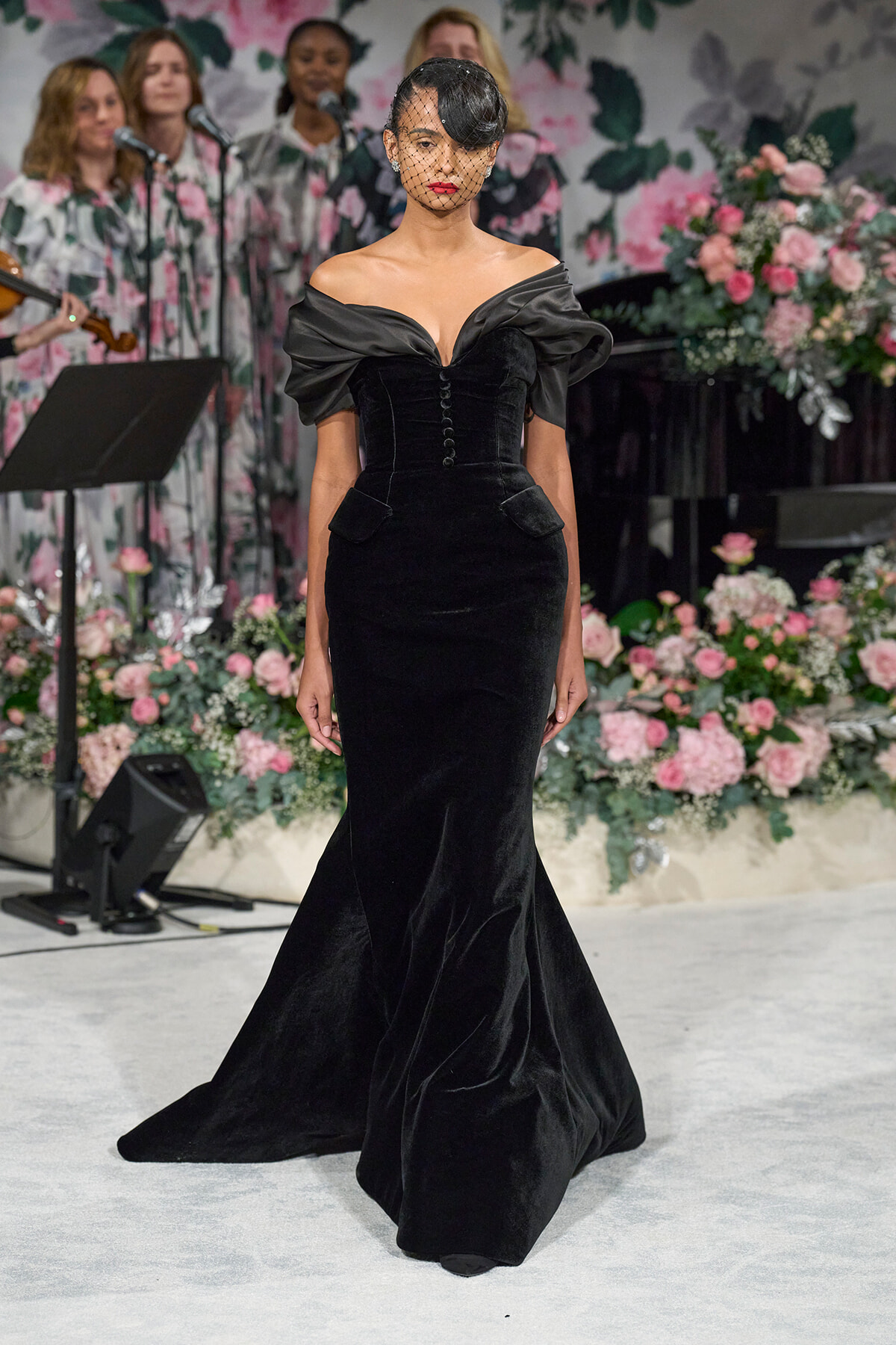Model walking the runway in a black off-the-shoulder velvet gown with a small veil over her face, floral backdrop behind her.
