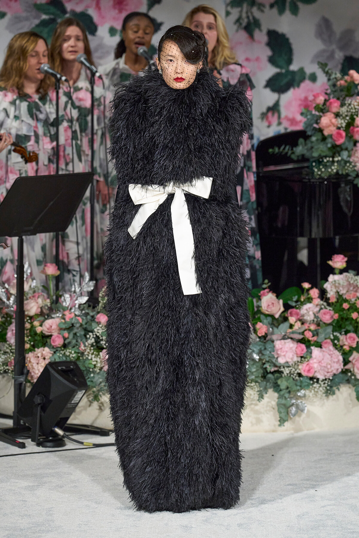Model in a black feathered gown with a white sash and veil on stage at a floral backdrop.