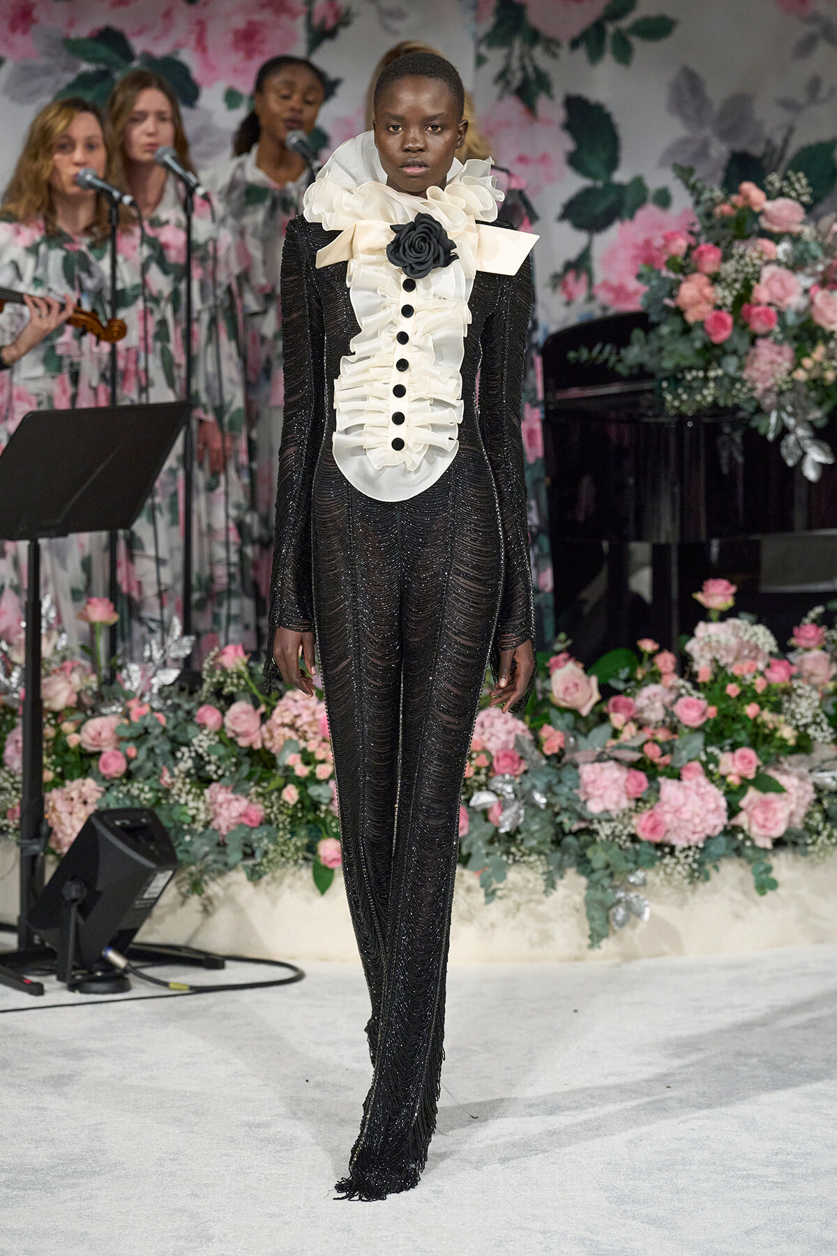Model in a black sequined gown with white ruffled bib and large black flower on the chest, standing on a floral runway stage.