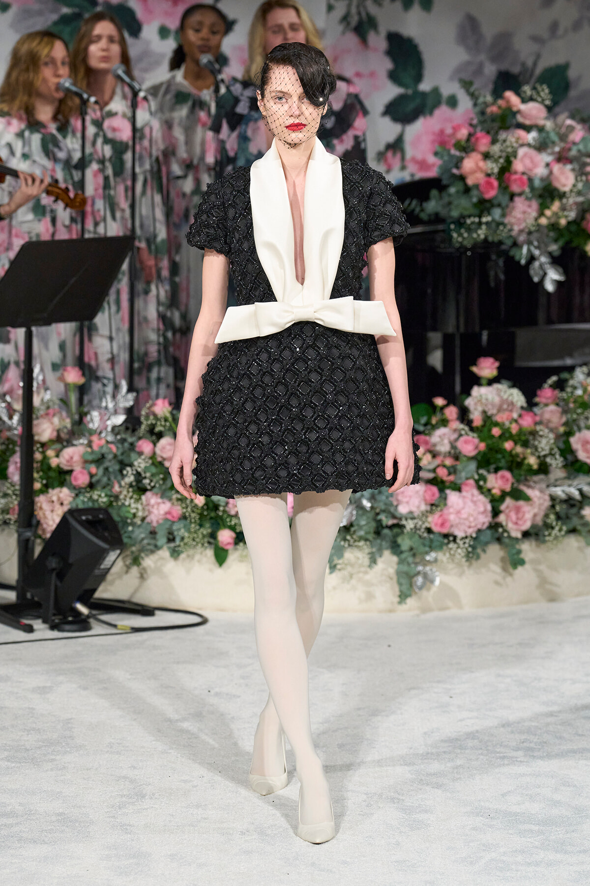 Model walking a fashion show wearing a black textured dress with a large white bow belt, white tights, and cream heels, her face partially veiled; floral backdrop and singers in the background.