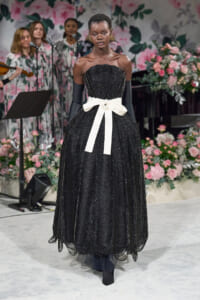 Model wearing a black beaded off-shoulder gown with a white sash walking a fashion show, floral backdrop in pinks and greens behind her.