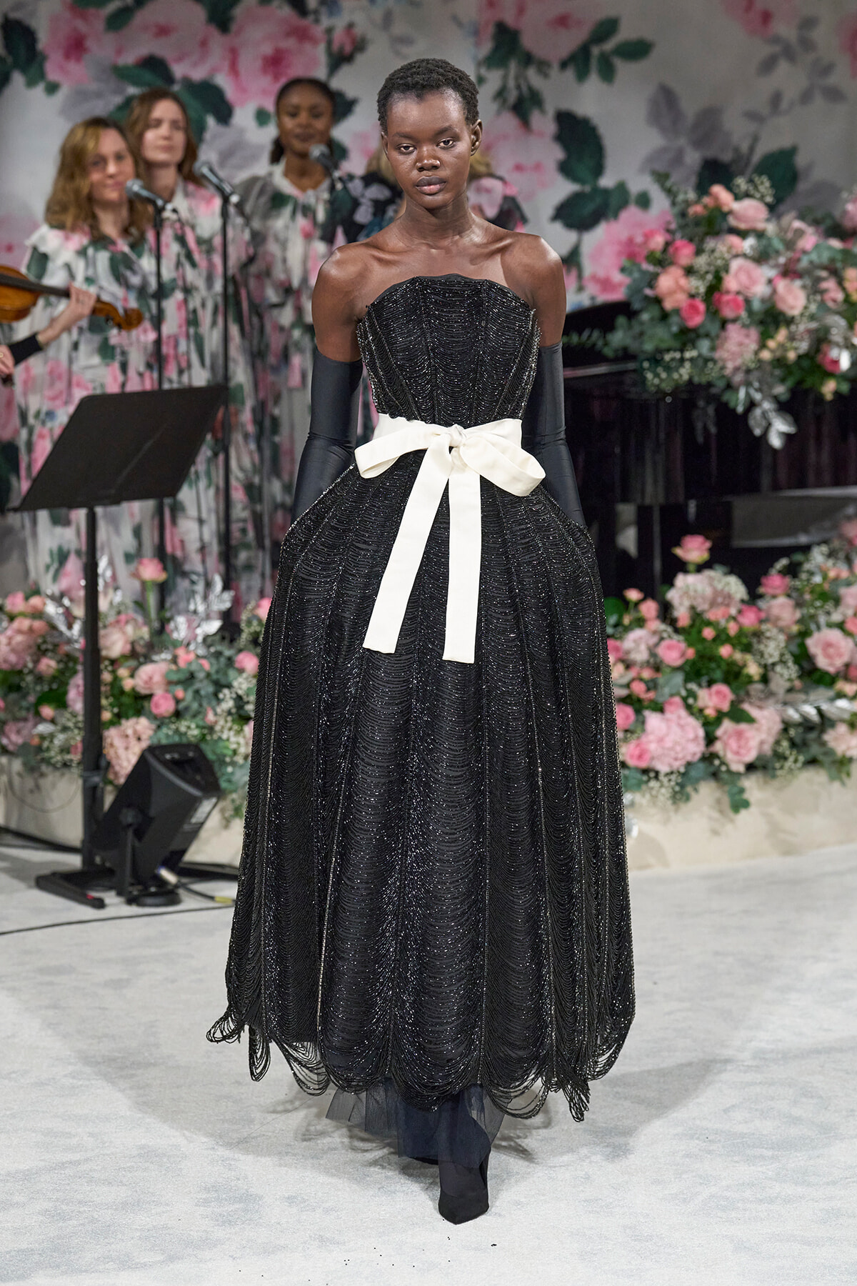 Model wearing a black beaded off-shoulder gown with a white sash walking a fashion show, floral backdrop in pinks and greens behind her.