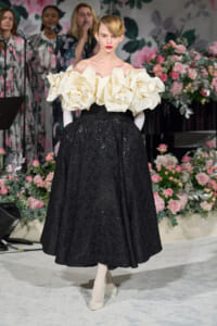 Model on a runway wearing a black lace ballgown with oversized cream ruffled off-shoulder detail, standing against a floral backdrop.
