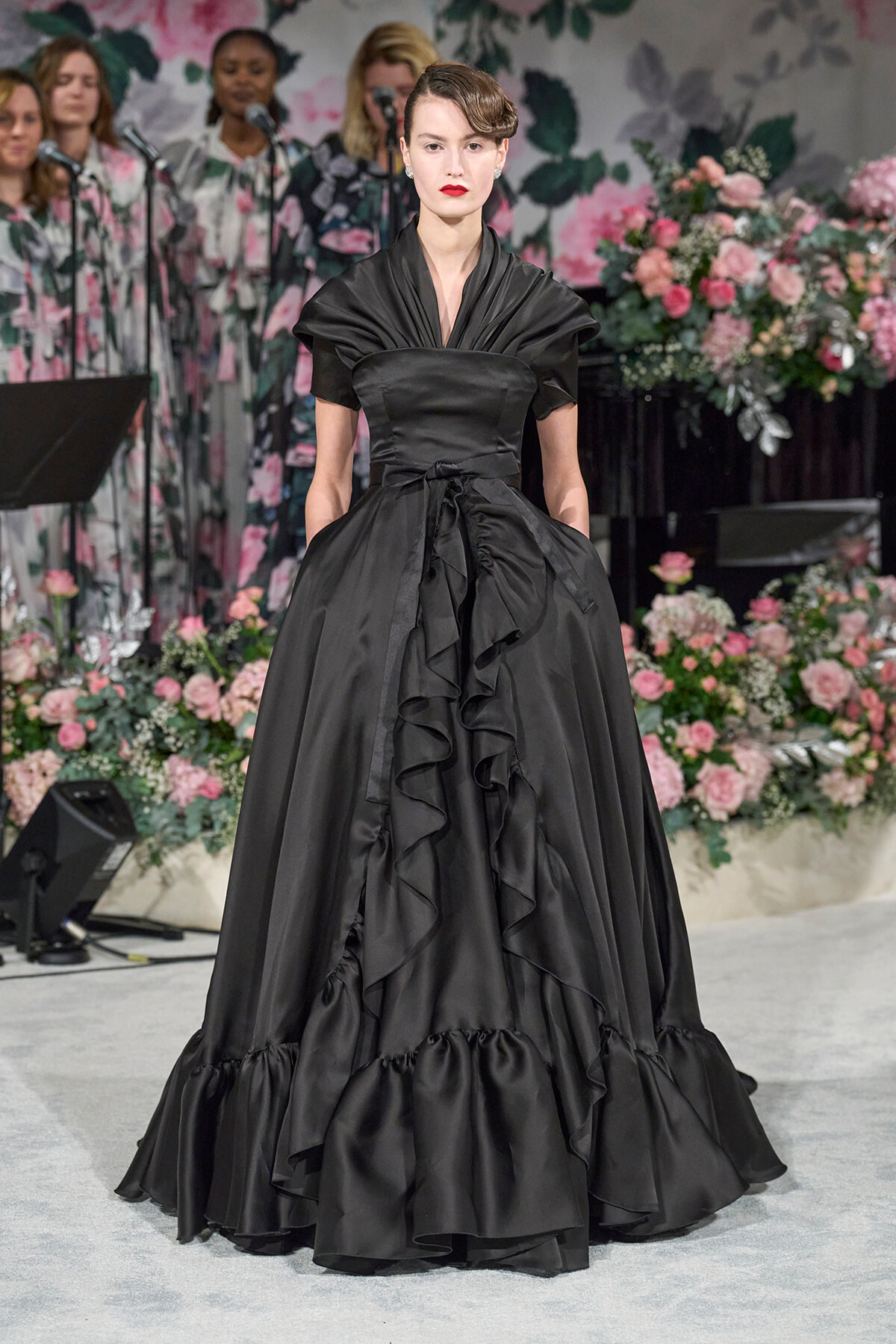 Model in a black satin ball gown with cascading ruffles on a runway, floral backdrop.