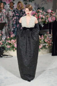 Model wearing a black sequined gown with a pearl-embellished white bodice and long black gloves on a floral runway backdrop
