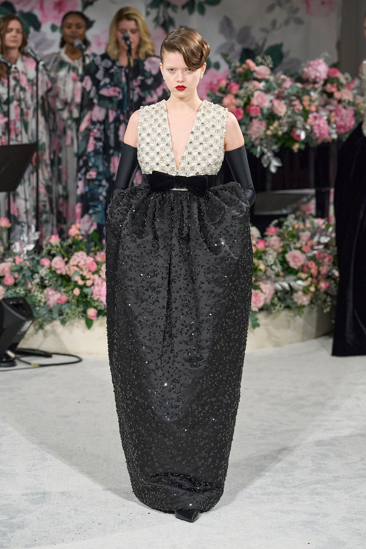 Model wearing a black sequined gown with a pearl-embellished white bodice and long black gloves on a floral runway backdrop