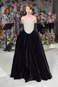 Model wearing a black velvet ball gown with a pearl-embellished bodice and long black gloves, standing on a floral-staged runway.