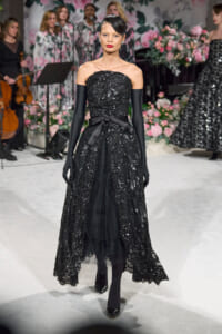 Female model in a black sequined off-the-shoulder gown with a satin bow belt and long gloves walking a fashion runway amid pink floral decor.