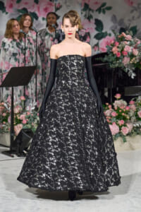 Model in a strapless black lace ballgown with long black gloves on a runway, with a pink floral backdrop behind her.