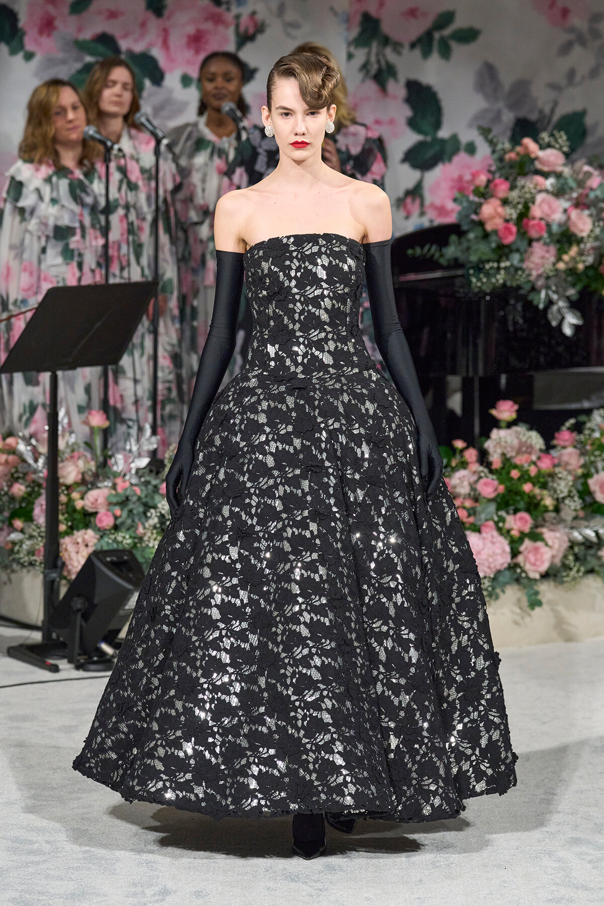 Model in a strapless black lace ballgown with long black gloves on a runway, with a pink floral backdrop behind her.