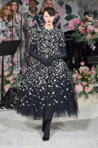 Model in a black, floor-length floral lace gown with sheer tulle hem and long black gloves, posing on a stage with a pink floral backdrop.