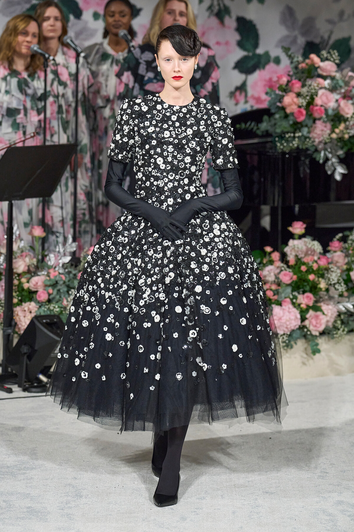 Model in a black, floor-length floral lace gown with sheer tulle hem and long black gloves, posing on a stage with a pink floral backdrop.