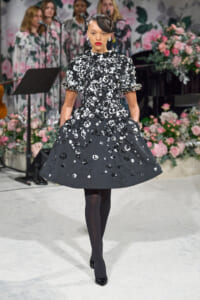 Model walking a runway in a black, knee-length dress covered in white floral appliqués, paired with black tights and patent heels, floral backdrop behind her