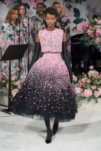 Model on a runway in a pink floral appliqué ball gown with long black gloves, against a backdrop of pink flowers and a singing group.