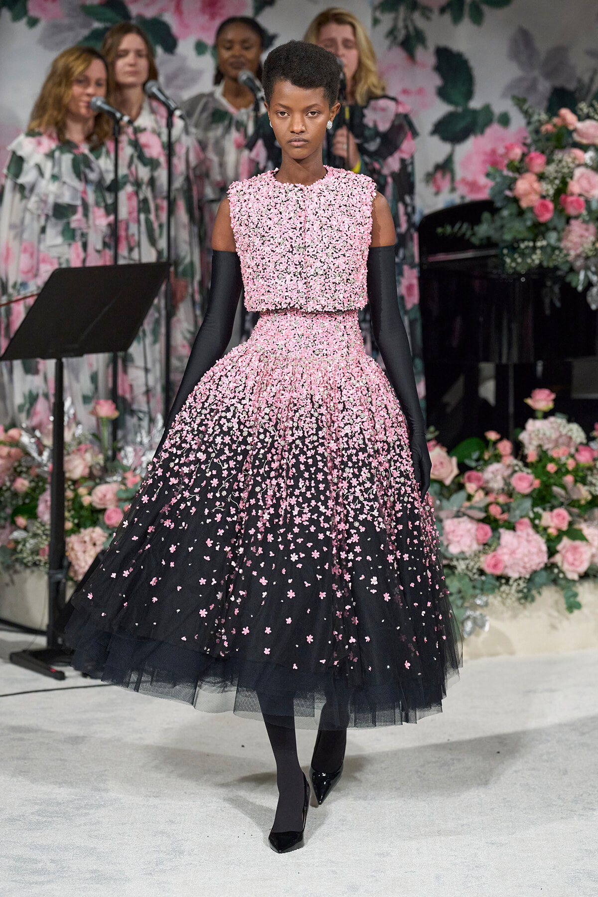 Model on a runway in a pink floral appliqué ball gown with long black gloves, against a backdrop of pink flowers and a singing group.