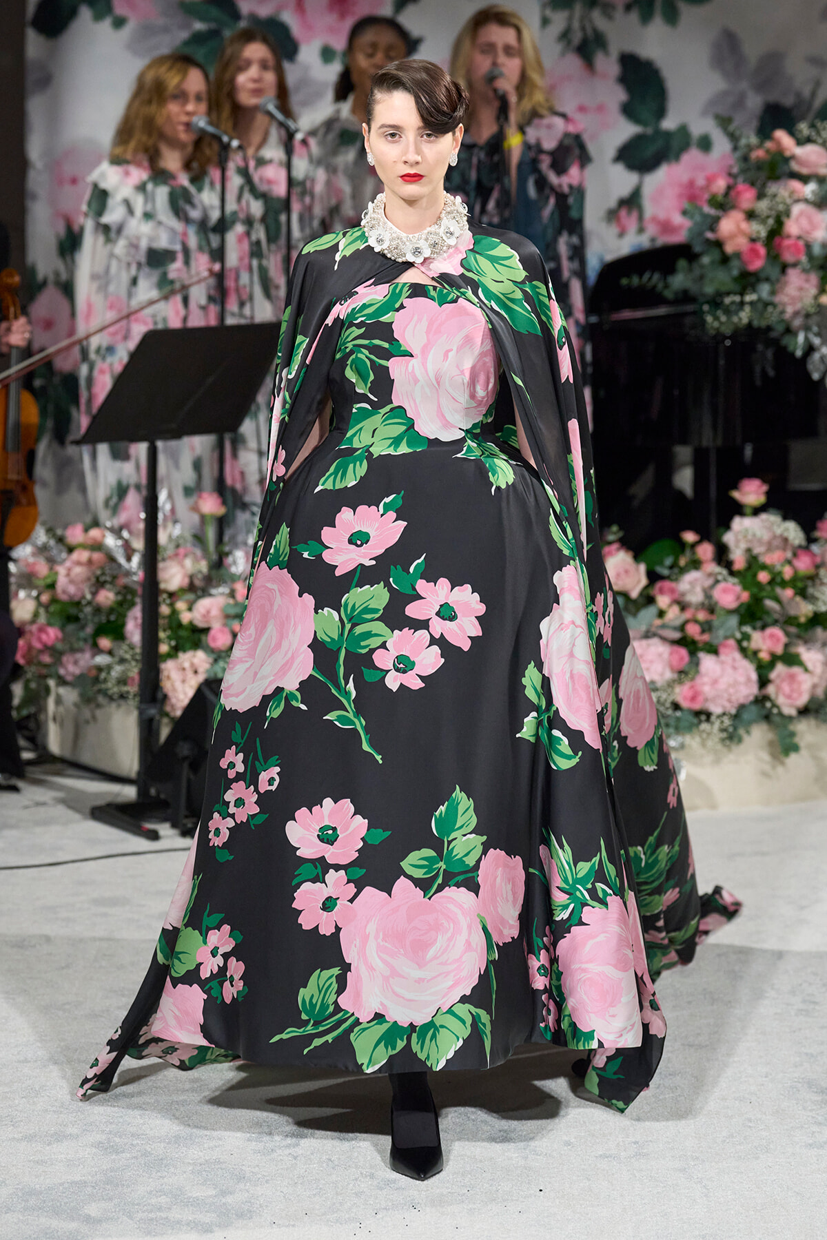 Model in a black gown with large pink floral print walks the runway, wearing a pearl necklace and bold makeup.