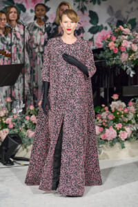 A woman in a pink floral-patterned gown with long black gloves stands on a flower-decorated stage, posing with one hand over her chest.