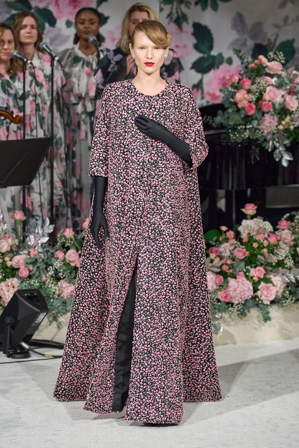 A woman in a pink floral-patterned gown with long black gloves stands on a flower-decorated stage, posing with one hand over her chest.
